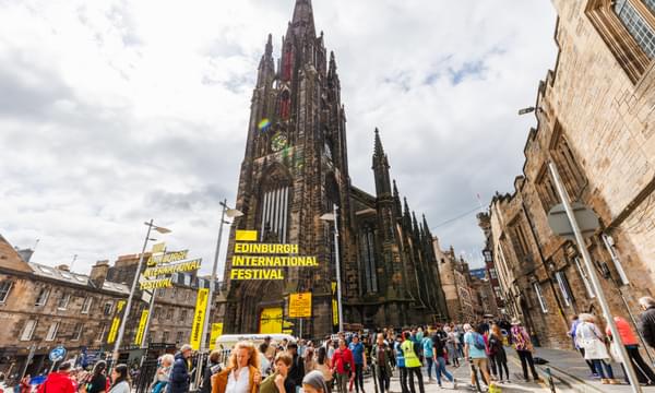 The Hub is surrounded by crowds of people, with a yellow sign reading 'Edinburgh International Festival' floating in front of the spire