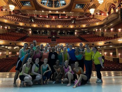 A group of 20 young people stand on stage smiling in a group photo. Their backs are facing the empty auditorium.