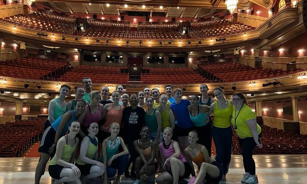 A group of 20 young people stand on stage smiling in a group photo. Their backs are facing the empty auditorium.