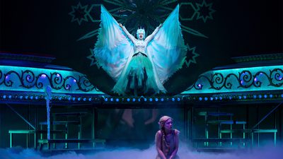 A women dressed entirely in white blue is stadning atop a podium which is lit with small blue lights. Her hands are above her head and her mouth is open wide. From her arms spread two shiney ice blue wings. She wears a blue crown on her head and a netted blue skirt. At the front of the image sits a small women who is hunched in fear. She has a blond braid on her shoulder.