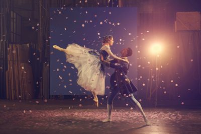 A women wearing a big skirt is being lifted up by a man dressed as a prince. They are both ballerinas and confetti falls around them.