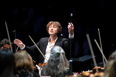A man stands wearning a suit in front of violin players. His arms are raised as he conducts the orchestra