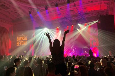 A person’s silhouette with their arms in the air and fingers spread in front of a brightly lit stage