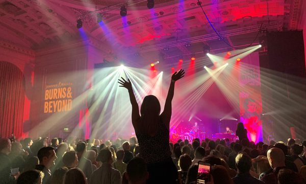 A person’s silhouette with their arms in the air and fingers spread in front of a brightly lit stage