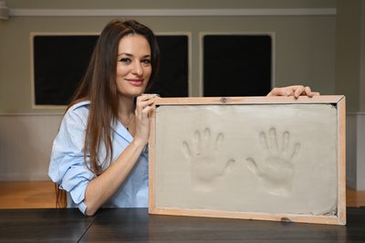 Nicola Benedetti, a woman with long brown hair, sits at a table with her elbow resting on it. She holds a clay model of her handprints slightly in front of her and is smiling. She is wearing a loose blue shirt and a black pendant necklace.
