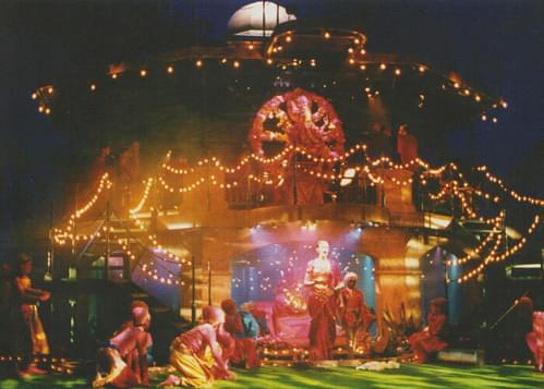 A brightly illuminated stage decorated with fairy lights and colourful canapes, with an actor standing centre stage dressed in a pink and gold sari.