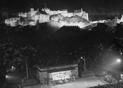 A black-and-white photo showing the Ross Bandstand in the foreground and Edinburgh Castle illuminated behind.