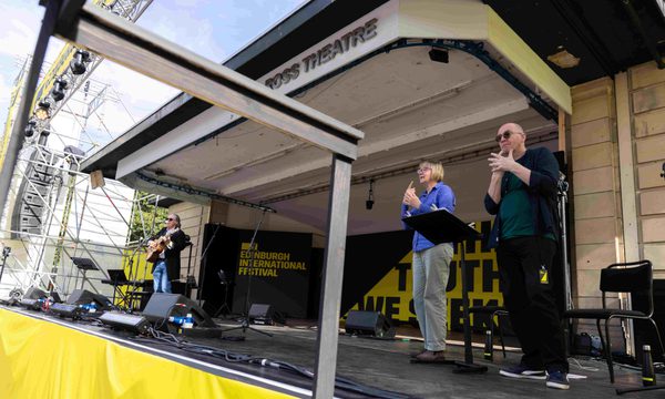 Two sign language interpreters stand on an outdoor stage. A man playing a guitar performs at centre stage.