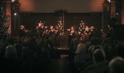 A group of string players are standing on stage. They are backlit by glowing candles. The audience sits in front of them in darkness