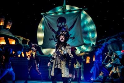 A man dressed in a gold and black long coat is standing on stage with his eyes and mouth wide. He has a hook instead of his left hand and a large feathered hat on his head. In the background, people dressed as pirates dance. There is a Skull and crossbones on a ships sail behind them.