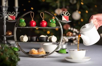 a white teapot is pouring black tea into a matching cup and saucer. In the background rests a 3-tiered serving platter with little christmas treats on it