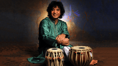 A man with shoulder length black curly hair is sitting in from of his tabla - an Indian drum. He is wearing a tunic and is smiling