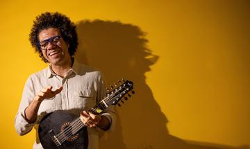 Man laughing holding stringed instrument, standing against yellow wall with bold shadows