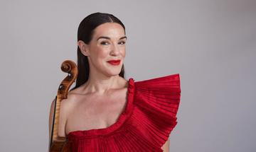 A dark-haired woman wearing a red dress smiles into the camera, a violin at her side