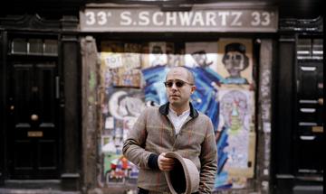 A man wearing dark sunglasses stands in front of a graffitied door holding a trilby in his hand
