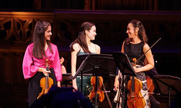 Three woman looking at each other smiling and holding stringed instruments