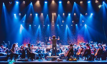 Orchestra performing on stage, conductor standing hands raised centre stage
