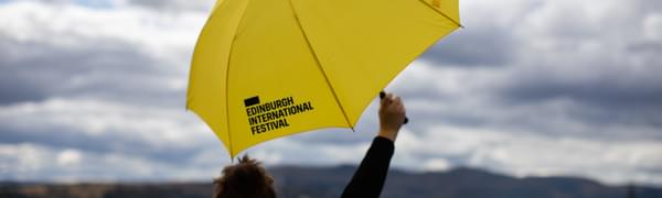 A yellow Edinburgh International Festival branded umbrella held aloft over the Edinburgh skyline by someone facing away from the camera.