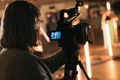 A woman stands looking into a screen on a camera as she films a live performance.