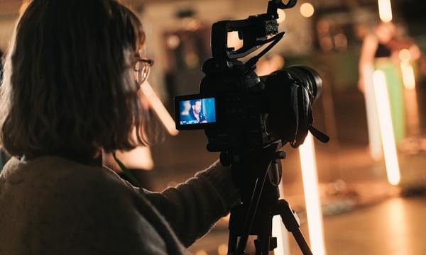 A woman stands looking into a screen on a camera as she films a live performance.