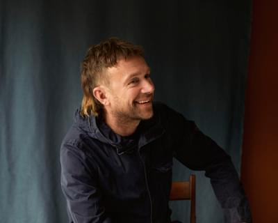 Damon Albarn sits on chair while smiling off to the side.