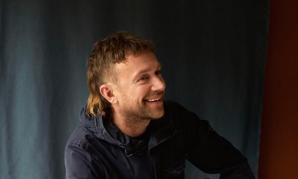 Damon Albarn sits on chair while smiling off to the side.