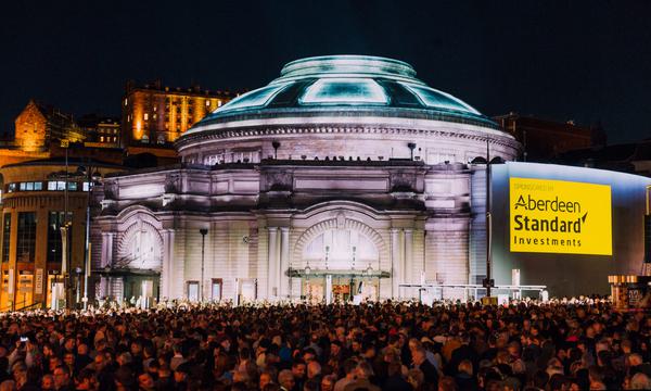 An illuminated Usher Hall with a crowd of people in front of him.