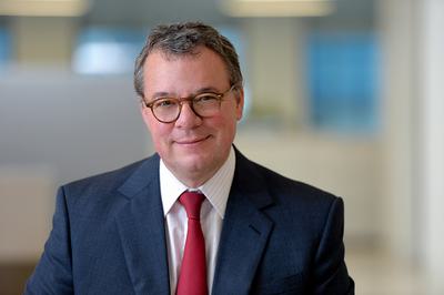 A man sits looking directly into the camera while wearing a navy blue suit, white shirt and red tie.