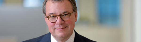 A man sits looking directly into the camera while wearing a navy blue suit, white shirt and red tie.
