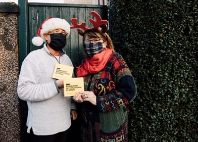 A couple group together outside while holding yellow Edinburgh International Festival CDs. They wear woolly jumpers, festive hats and masks.