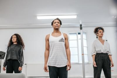 Courtney Stoddart, Patricia Panther and Saskia Ashdown stand in a socially-distanced line within a bright rehearsal room, staring out beyond the photographer’s camera. Each have their arms by their sides and all wear monochromatic clothing.