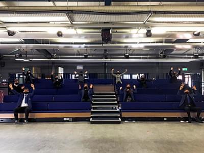 Students sit socially distanced on a blue raised seating bank while waving their hands in the air.