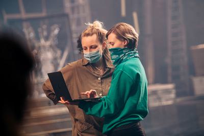 Choreographers Jess & Morgs stand masked up backstage at a theatre both starind down into a black laptop.