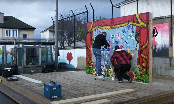 Two men stand on an outdoor stage while installing a colourful backdrop.