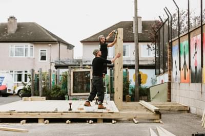 Two men stand in a playground installing a wooden stage.