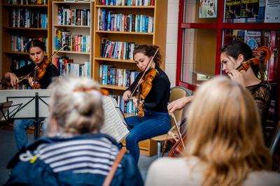 Three people sit on chairs playing different string instruments as a group of people watch their performance.