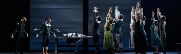 A scene from Scottish Ballet's production of The Crucible showing a group of people facing towards the back of the stage, their arms above their heads. A young girl lies on a table in the background.