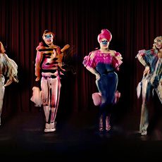 An image depicting The Zizi Show, a deepfake interactive drag cabaret. Six deepfake drag artists stand in a line in colourful costume while striking a variety of poses against a dark red curtain backdrop.