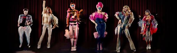 An image depicting The Zizi Show, a deepfake interactive drag cabaret. Six deepfake drag artists stand in a line in colourful costume while striking a variety of poses against a dark red curtain backdrop.