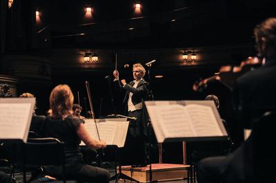 A conductor stands facing his orchestra while wearing a tux and holding his baton in the air. In front of him is a selection of string players reading sheet music from stands.