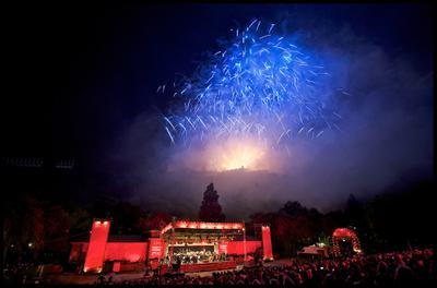 Blue fireworks illuminate the sky as an orchestra plays on a red stage below them.