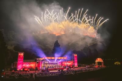 Fireworks illuminate the sky above a crowd watching an orchestra perform on a red stage.
