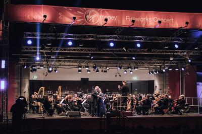 An orchestra performs on stage led by a conductor as two singers embrace in front of them.