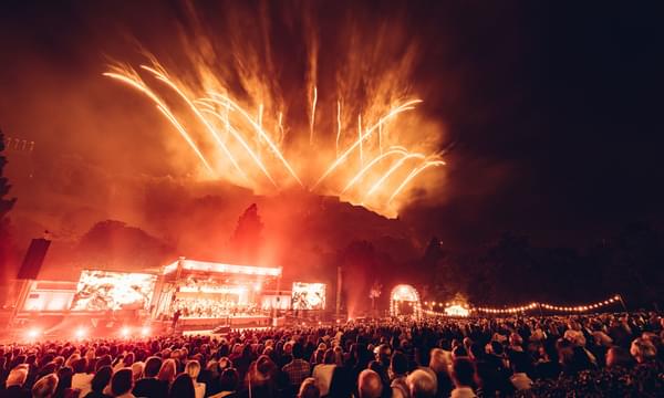 Red fireworks fill the sky as red lighting illuminates a stage in Princes Street Gardens. A crowd watches on as an orchestra plays on the stage.