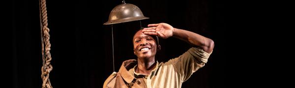 A man in army uniform smiles and salutes, standing beneath a suspended steel helmet.