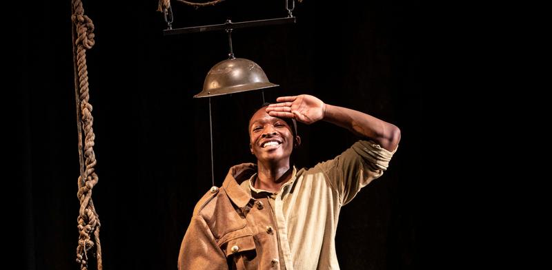 A man in army uniform smiles and salutes, standing beneath a suspended steel helmet.