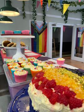 A selection of rainbow inspired bakes are displayed in a room decorated with progress flags and rainbow bunting