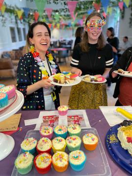 Two members of EIF staff stand smiling wearing rainbow flowers and Pride glasses holding plates full of rainbow cake