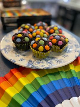 A plate of chocolate cupcakes decorated with rainbow smarties on top, sat next to a rainbow fan