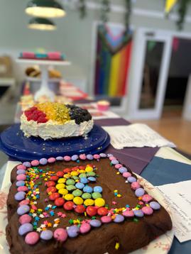 A selection of rainbow inspired bakes are displayed in a room decorated with progress flags and rainbow bunting. In the foreground are brownies with a rainbow of smarties on top.